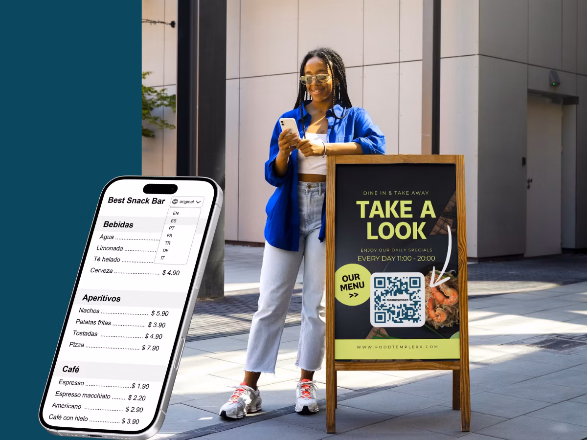 Smiling small restaurant owner standing in front of two digital momoscreens showing food and drink menus in a casual kitchen setting.